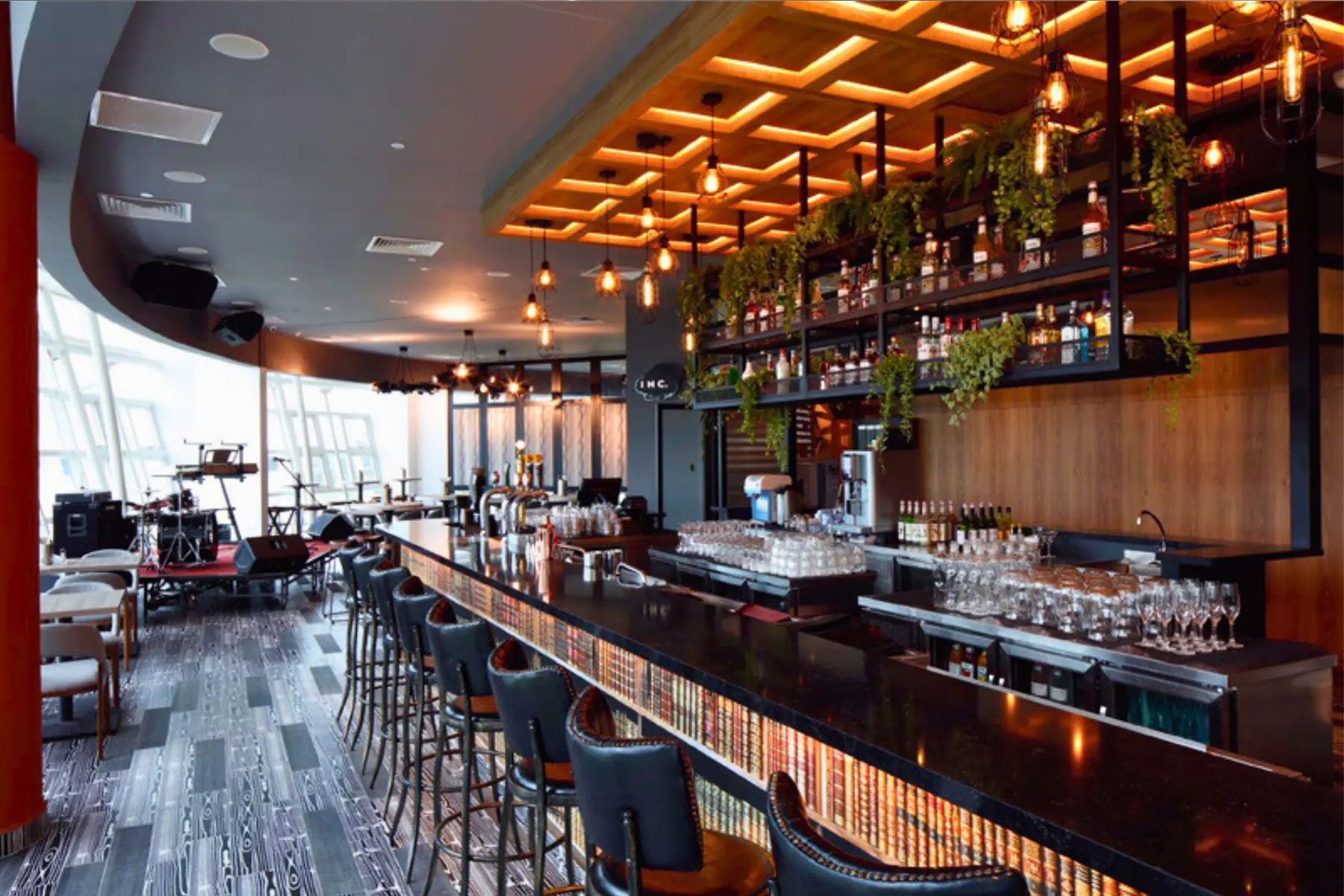 The long bar at PEN & INC NTU, backed by a lit ceiling grid and spirit shelves, with the live stage by the curved window wall